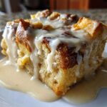 A close-up of a square slice of rich bread pudding generously topped with a thick vanilla cream sauce.