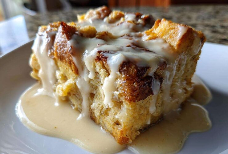 A close-up of a square slice of rich bread pudding generously topped with a thick vanilla cream sauce.