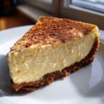 A creamy slice of eggnog cheesecake with a dark graham cracker crust, dusted with cocoa powder.