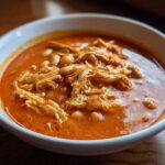 Close-up of a white bowl filled with vibrant orange buffalo chicken chili, featuring shredded chicken and white beans.