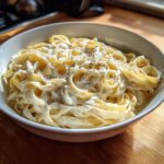 A white bowl filled with fettuccine pasta generously coated in creamy homemade alfredo sauce and topped with black pepper.