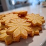 Close-up of golden, flower-shaped butter cookies stacked on a white plate, sprinkled with sugar.