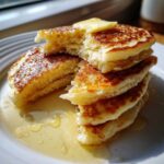 A stack of fluffy buttermilk pancakes cut open, topped with melting butter and drizzled with syrup.