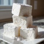 A stack of four large, square homemade marshmallows dusted heavily with powdered sugar on a white plate.