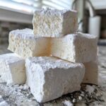 A stack of six fluffy, square homemade marshmallows heavily dusted with powdered sugar on a granite countertop.