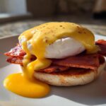 A single serving of eggs benedict featuring a toasted English muffin, Canadian bacon, a poached egg, and rich hollandaise sauce dripping onto the plate.