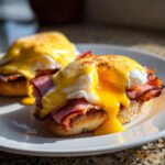 Two servings of classic eggs benedict on English muffins with ham, topped with rich hollandaise sauce, yolk running out.