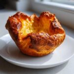 A single, tall, golden-brown yorkshire pudding sitting on a white plate, showcasing its high rise.