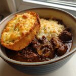 A bowl of rich French onion beef stew topped with melted cheese and a slice of cheesy garlic bread.