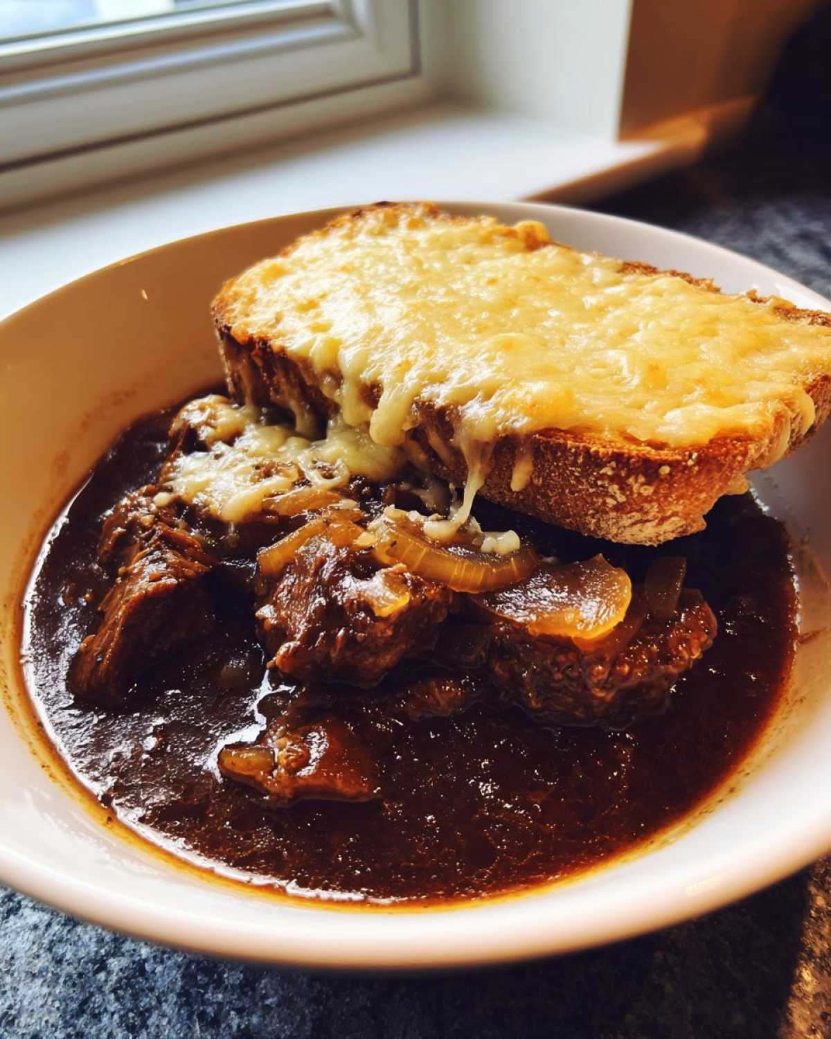 A bowl of rich French onion beef stew topped with tender beef chunks and a slice of bread covered in melted cheese.
