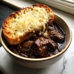A close-up of rich French onion beef stew topped with a large, cheesy, broiled crouton.