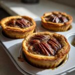 Close-up of three freshly baked mini pecan pies cooling in a light gray muffin tin.