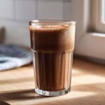 A tall, fluted glass filled with rich, frothy chocolate milk sitting on a sunlit wooden countertop.