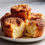 Close-up of several golden brown french toast muffins topped with cinnamon sugar, one is broken open.