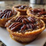 Four glistening mini pecan pies with caramelized filling and whole pecans on top, served on a white rectangular plate.