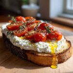 Close-up of gourmet ricotta toast topped with roasted cherry tomatoes, thyme, and olive oil drizzling down the crust.