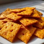 A pile of bright orange, square homemade cheese crackers with small holes, displayed on a white plate.
