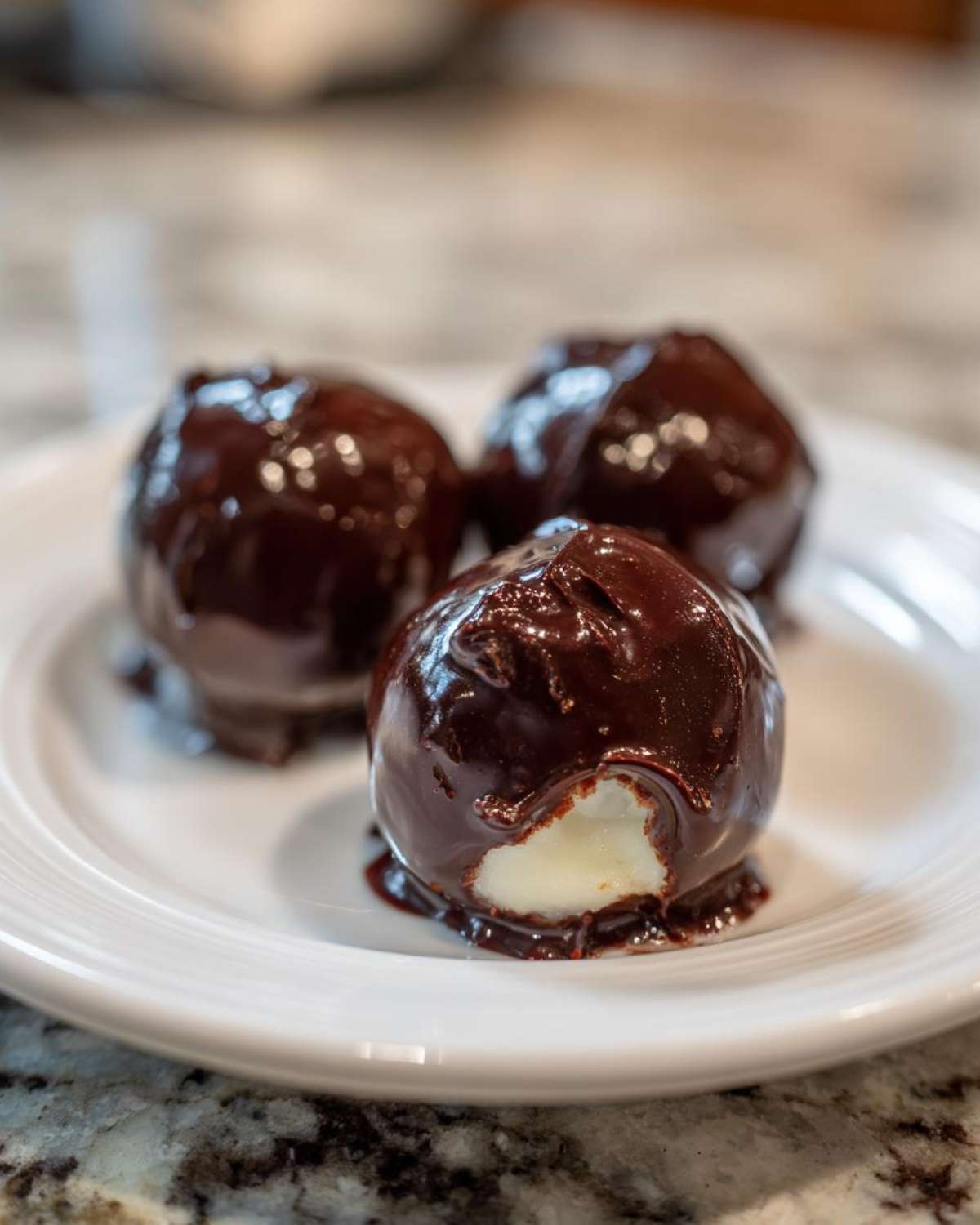 Three homemade chocolate covered cherries on a white plate, one with a bite showing the creamy white filling.