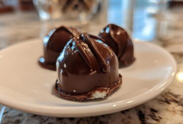 Close-up of three homemade chocolate covered cherries resting on a white plate, showing the glossy dark chocolate coating.