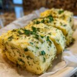 Three thick slices of homemade garlic butter mixed with green herbs, resting on parchment paper.