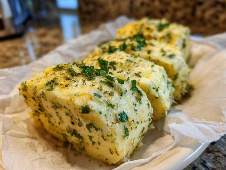 Three thick slices of homemade garlic butter mixed with green herbs, resting on parchment paper.