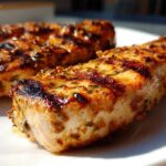 Close-up of two perfectly grilled chicken breasts coated in a rich, herb-flecked chicken marinade, showing char marks.