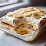 A glass dish containing a layered, creamy banana pudding dessert topped with whipped cream and fresh banana slices.