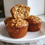 Three golden brown banana oatmeal muffins stacked on a white plate, one cut open showing the moist oat texture.