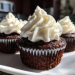 Three moist gingerbread cupcakes topped with swirls of thick white cream cheese frosting, presented on a white plate.