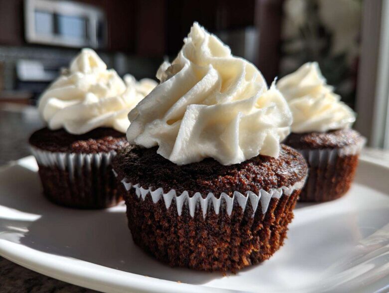 Three moist gingerbread cupcakes topped with swirls of thick white cream cheese frosting, presented on a white plate.