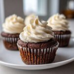 A close-up of a moist gingerbread cupcake topped with a swirl of cream cheese frosting, with other gingerbread cupcakes blurred in the background.