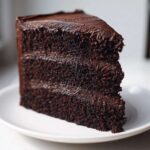A close-up of a tall, moist slice of homemade chocolate cake layered with dark chocolate frosting on a white plate.