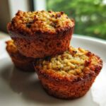 Three golden-brown, moist oatmeal muffins stacked on a white plate with a blurred green background.