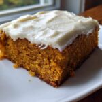 A close-up of a moist, square slice of pumpkin bars with thick cream cheese frosting on a white plate.