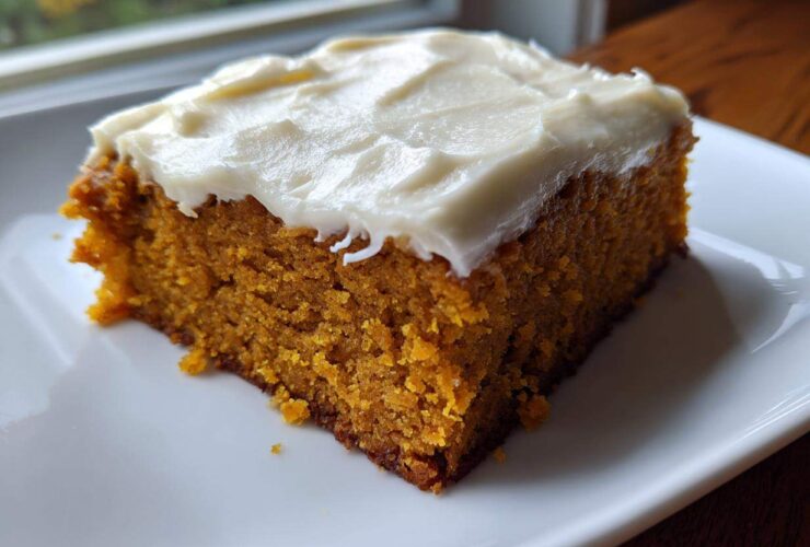 A close-up of a moist, square slice of pumpkin bars with thick cream cheese frosting on a white plate.