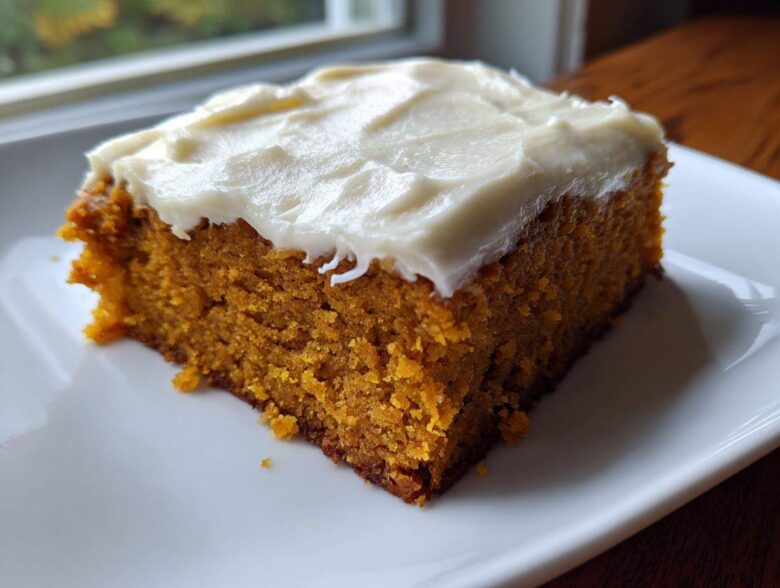 A close-up of a moist, square slice of pumpkin bars with thick cream cheese frosting on a white plate.