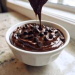 A white bowl filled with thick, glossy brownie batter dip, topped with chocolate chips, as more dip is drizzled on top.