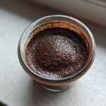 Overhead close-up of rich homemade chocolate pudding dusted with cocoa powder in a small glass jar.