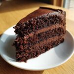 A moist, dark slice of chocolate cake recipe with thick layers of frosting on a white plate.