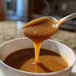 Close-up of rich, brown turkey gravy steaming as it drips from a spoon back into a white bowl.
