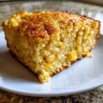 Close-up of a moist slice of cornbread casserole showing golden brown top and visible yellow corn kernels throughout.
