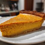 A close-up of a perfect slice of rich, orange vegan pumpkin pie on a white plate.