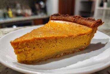 A close-up of a perfect slice of rich, orange vegan pumpkin pie on a white plate.