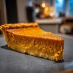 A close-up, appetizing slice of rich orange vegan pumpkin pie resting on a dark plate.