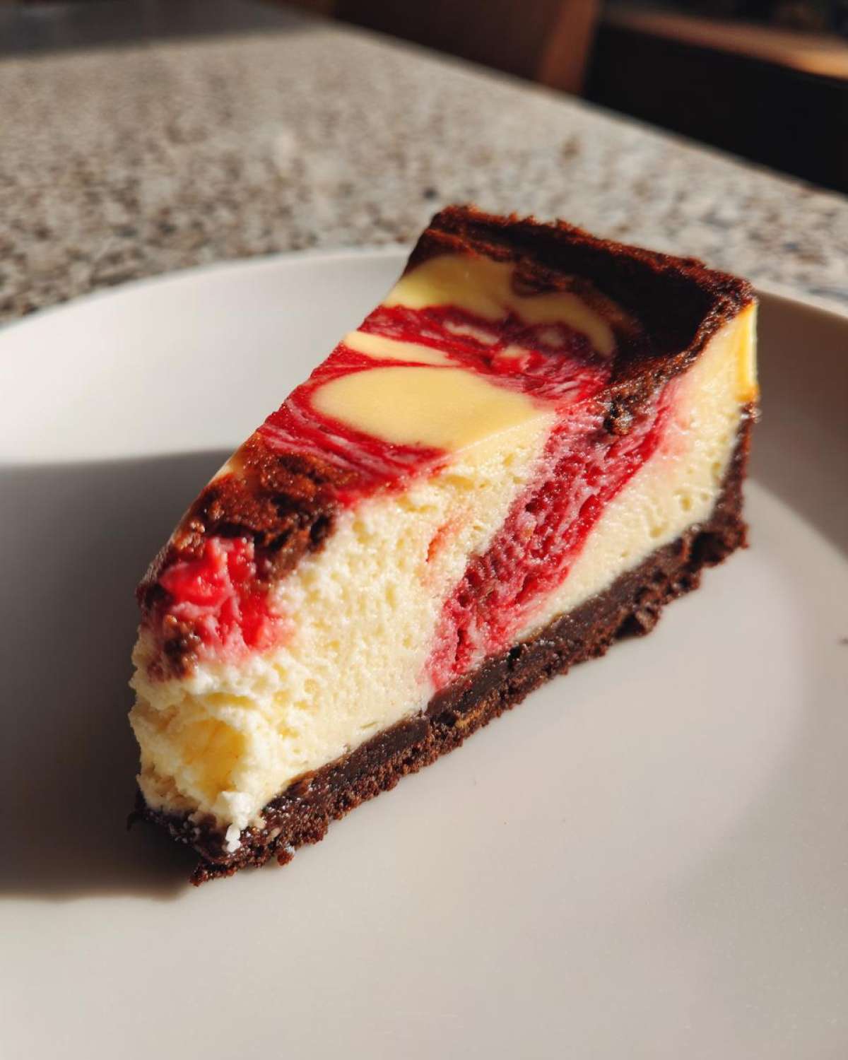 A single slice of white chocolate raspberry cheesecake featuring a dark crust and a creamy filling swirled with bright red raspberry.