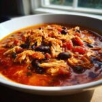 A close-up, appetizing view of shredded chicken, black beans, and tomatoes in a rich red broth, representing slow cooker buffalo chicken chili.