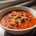 A white bowl filled with vibrant red slow cooker buffalo chicken chili, featuring shredded chicken and black beans.