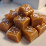 A close-up stack of soft, chewy, golden-brown caramels topped with flaky sea salt.