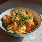 A close-up of a bowl of spicy kani salad, featuring shredded crab mixed with orange dressing, topped with sesame seeds and green onions.