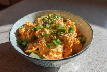 A close-up of a bowl of spicy kani salad, featuring shredded crab mixed with orange dressing, topped with sesame seeds and green onions.